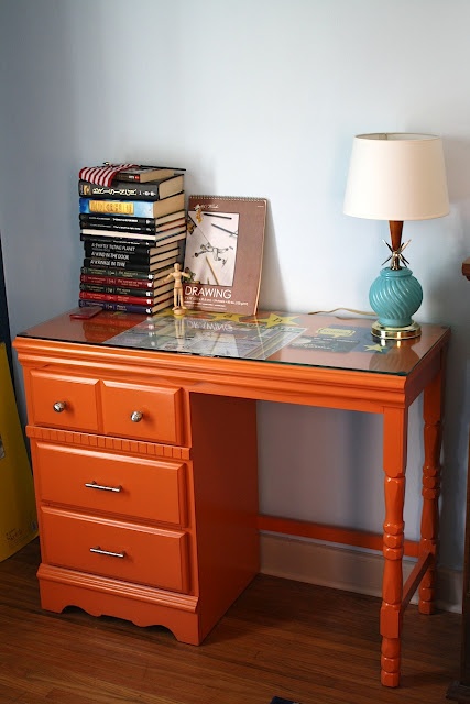 an orange desk with two drawers and a lamp on it next to a stack of books