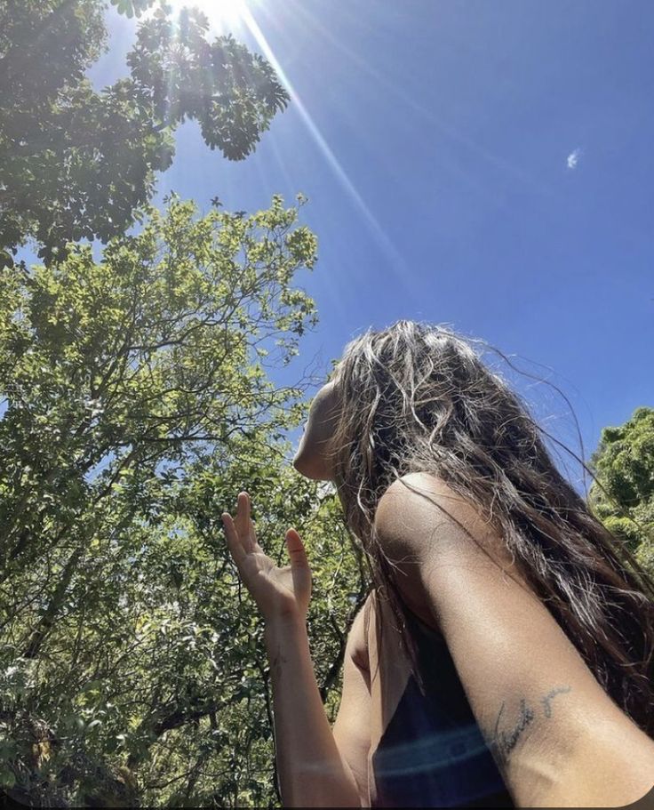 a woman standing in front of trees with her hand up to the sky and sun shining on her face