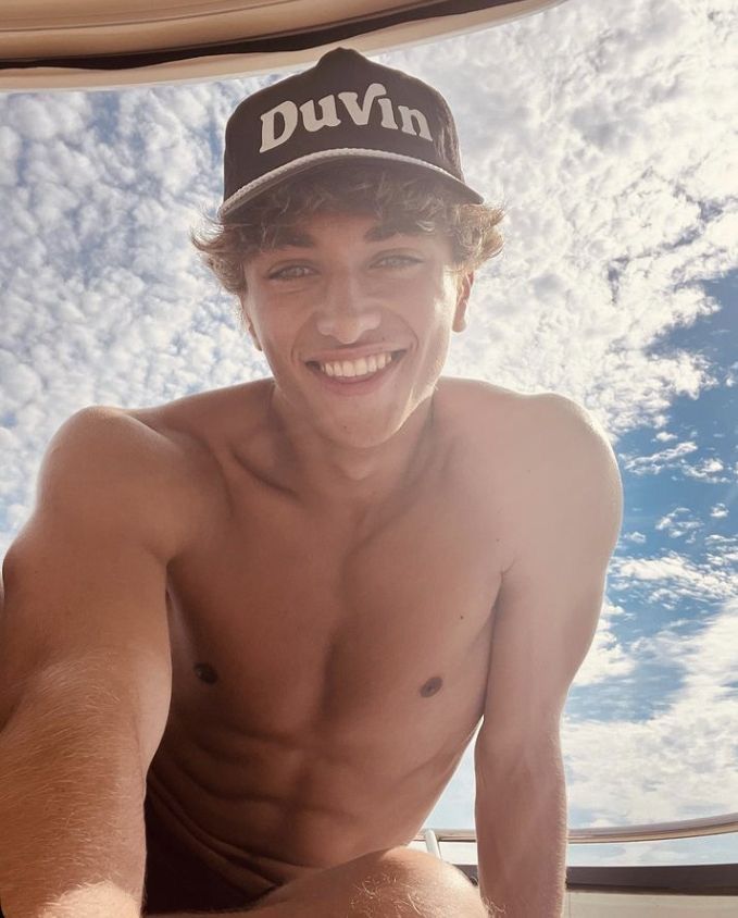 a shirtless young man sitting on the back of a boat with his hat over his head