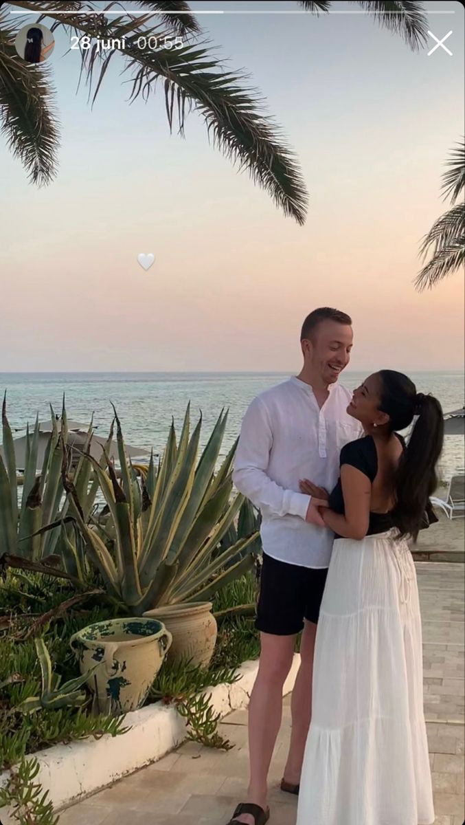 a man and woman standing next to each other in front of the ocean with palm trees