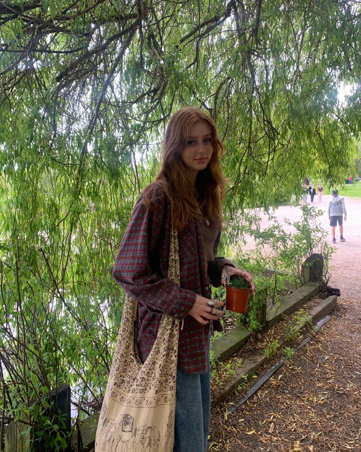 a woman standing under a tree with a bag and bucket in her hand while holding a cup