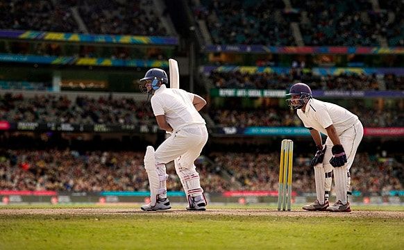 two men in white uniforms playing a game of cricket on a field with people watching from the stands