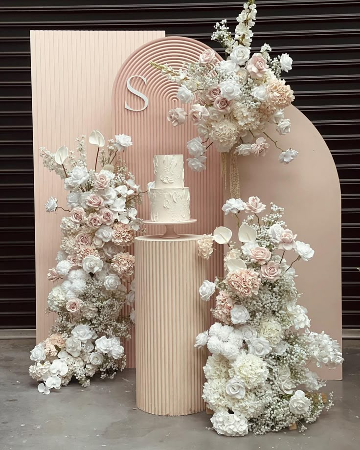 a wedding cake surrounded by flowers and greenery in front of a pink backdrop with an arch