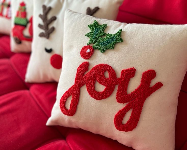 three christmas pillows with the word joy embroidered on them sitting on a red couch cushion