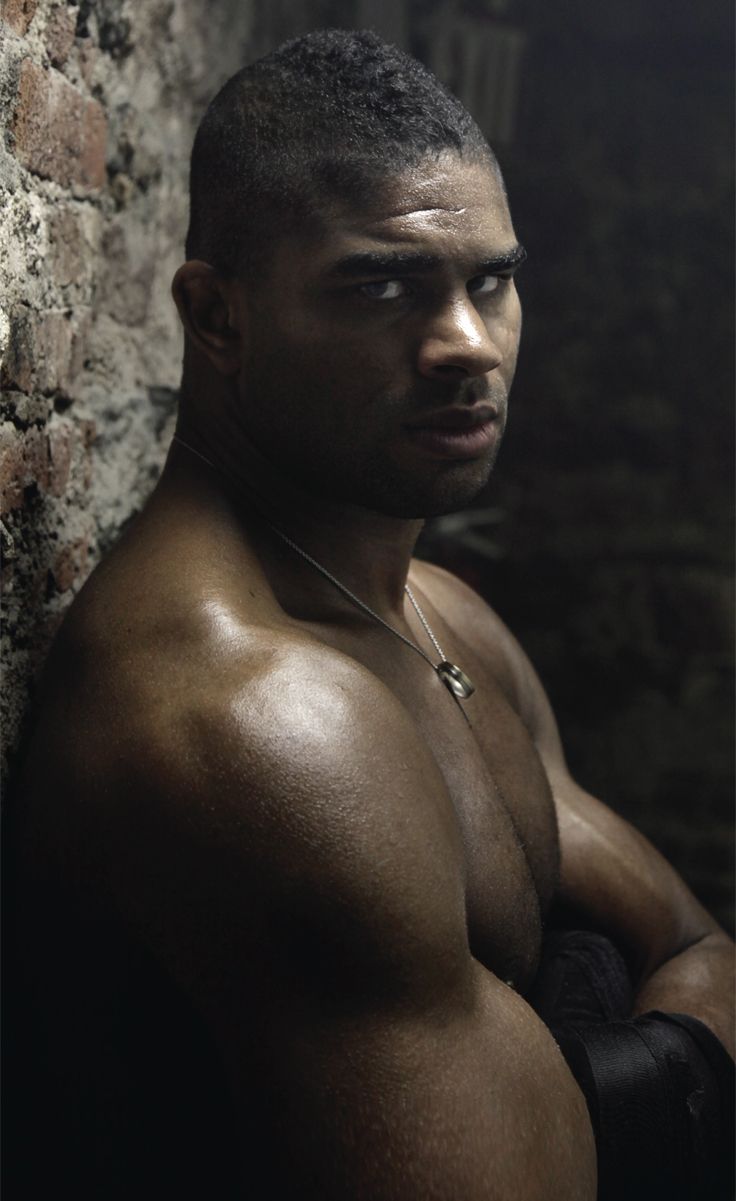 a shirtless man leaning against a brick wall with his hands on his hips looking at the camera