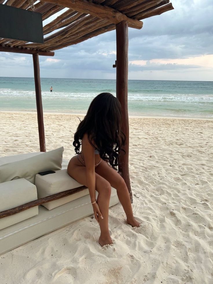 a woman sitting on top of a white couch under a wooden structure next to the ocean