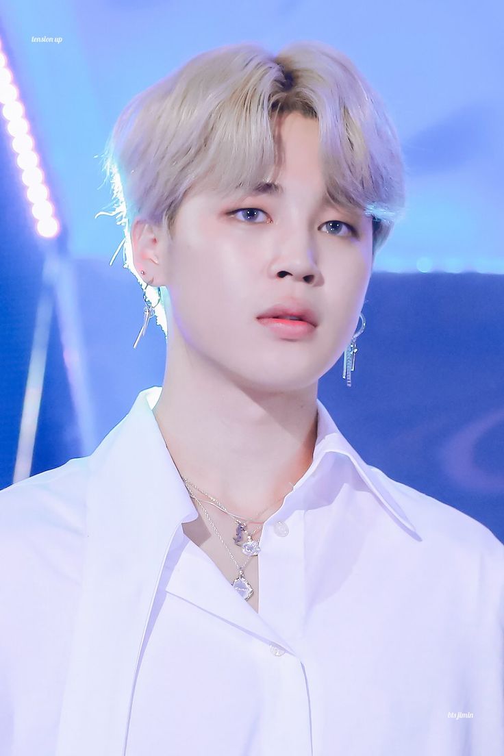 a male in a white shirt and silver necklace on a blue background with lights behind him