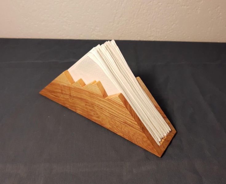 a wooden triangle with folded papers in it on a black tableclothed surface next to a white wall