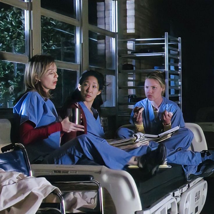 three women in scrubs are sitting on a hospital bed and talking to each other