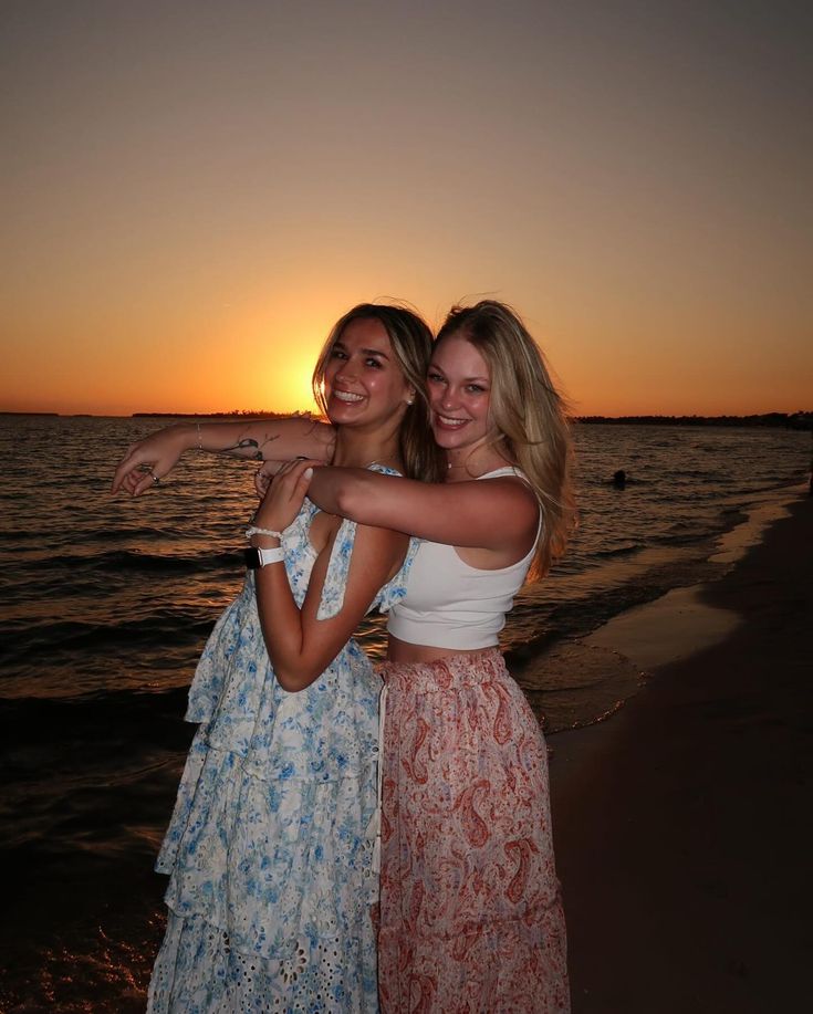 two women standing next to each other on a beach near the ocean at sunset with their arms around each other