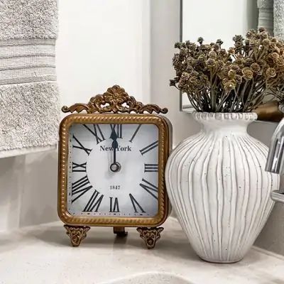 a clock sitting next to a white vase with dried flowers in it on a bathroom counter