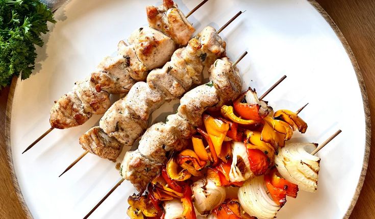 a white plate topped with meat and veggies on skewers next to parsley