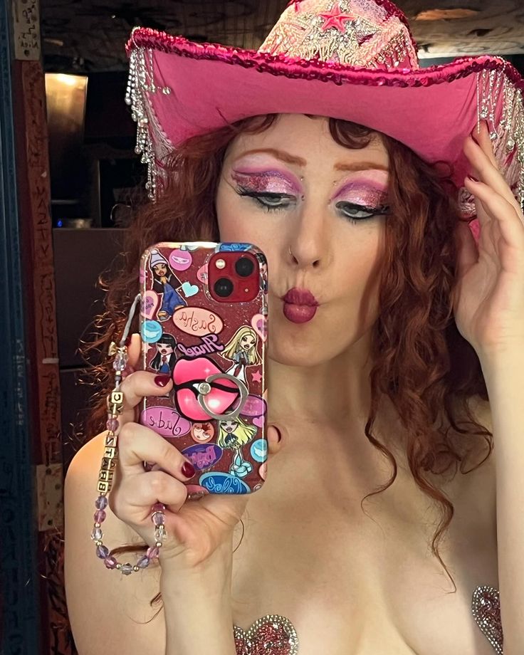 a woman with red hair wearing a pink cowboy hat and holding a cell phone up to her face