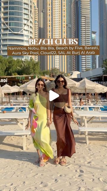 two women standing on top of a beach next to the ocean and tall buildings in the background