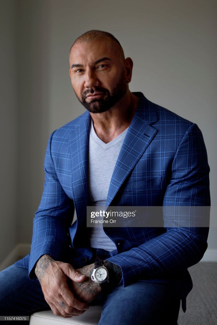 a bald man in a blue suit sits on a stool and looks off to the side