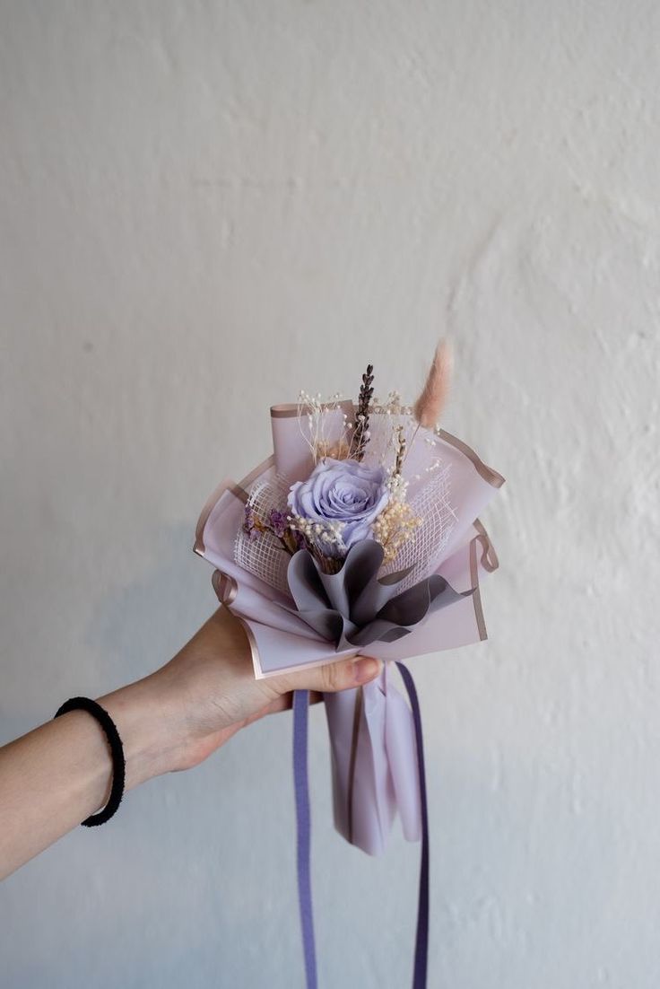 a hand holding a paper flower with purple ribbon and beading on the end, in front of a white wall