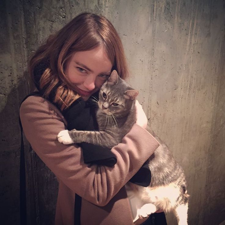 a woman holding a cat in her arms while standing next to a wall and looking at the camera