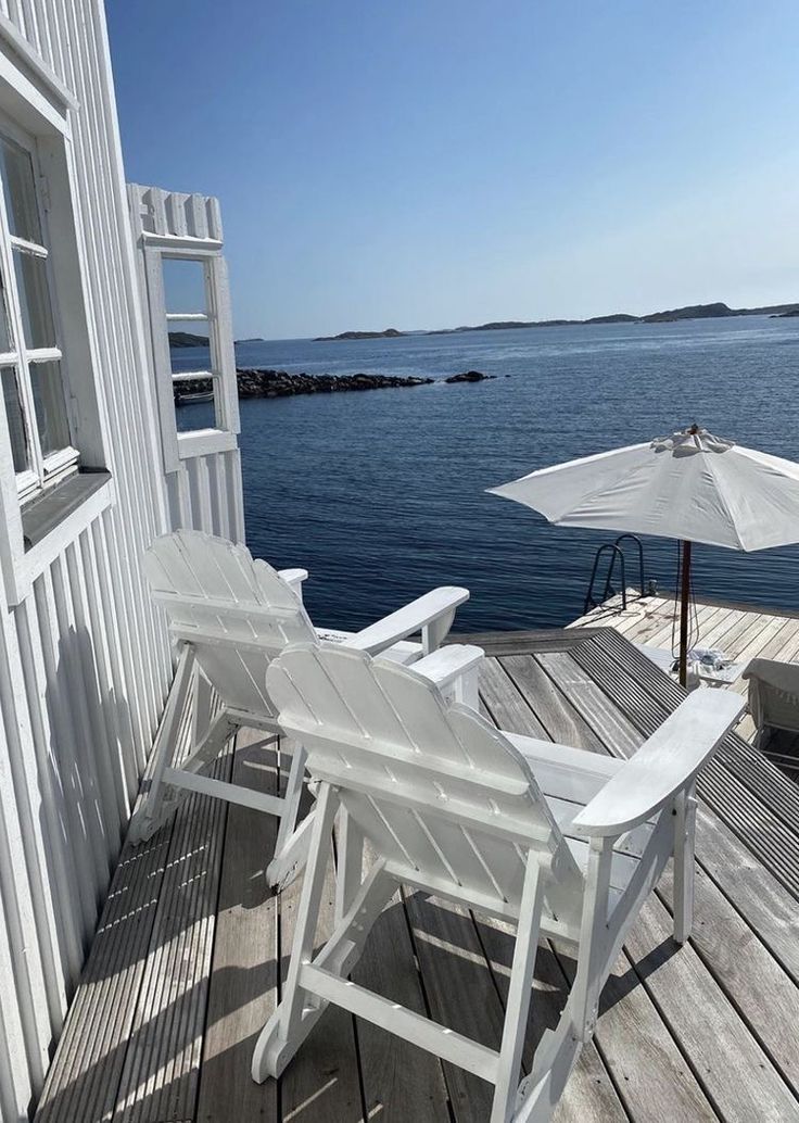 two white lawn chairs sitting on top of a wooden deck next to an umbrella and water