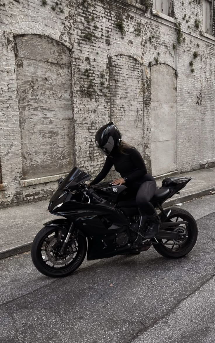 a woman in black riding on the back of a motorcycle down a street next to a brick building