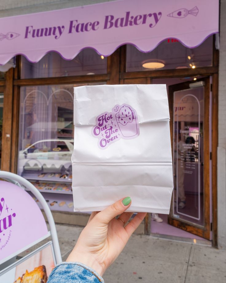 a person holding up a paper bag in front of a pastry shop with purple awnings