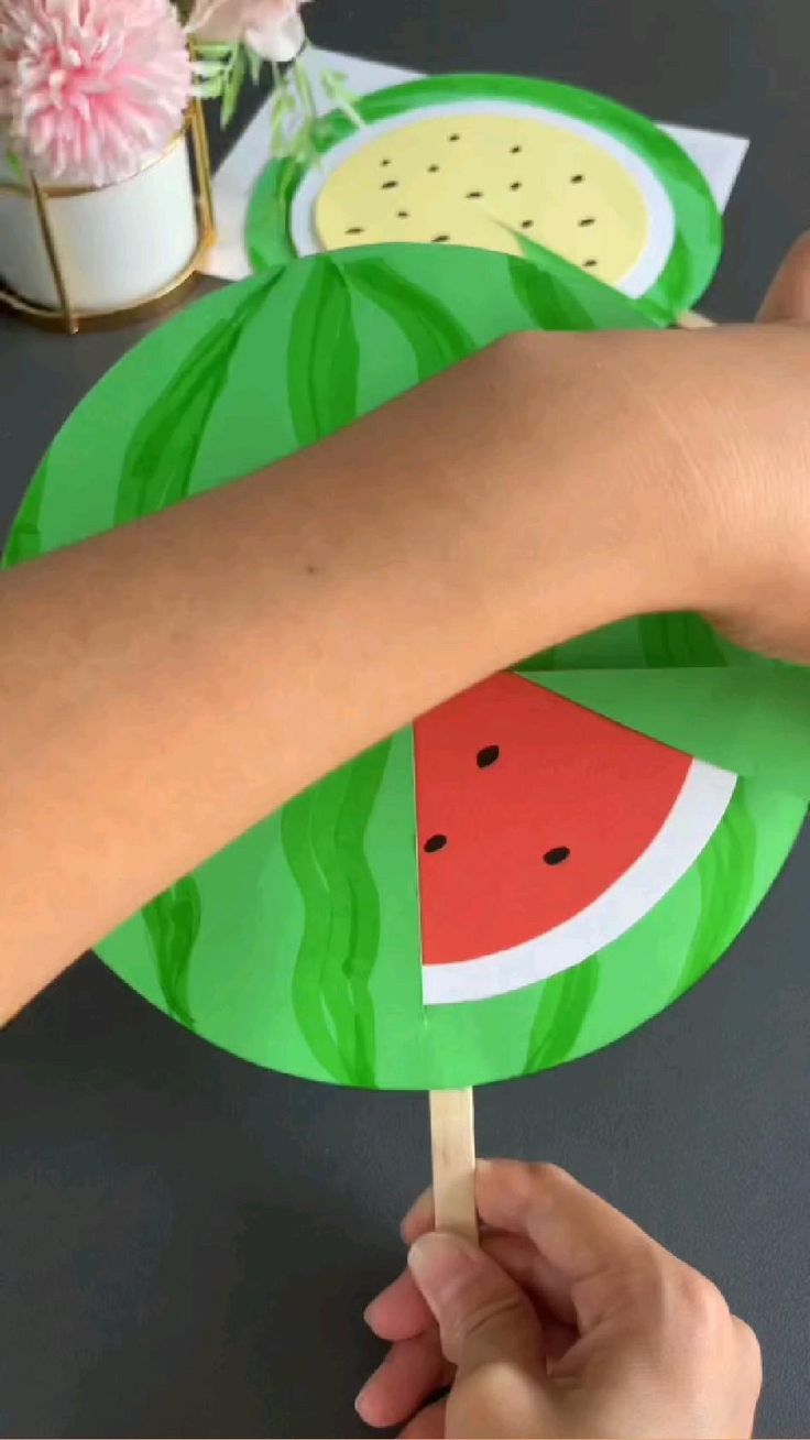 a person is holding a paper plate with watermelon slices on it while another hand holds a stick