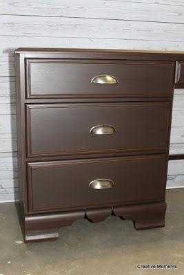 a brown dresser with three drawers and two doors on the bottom drawer, in front of a white wall