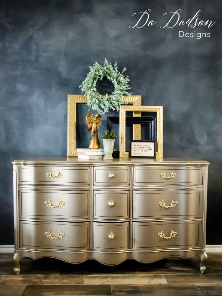 a gold dresser with two framed pictures and a wreath on top, in front of a chalkboard wall