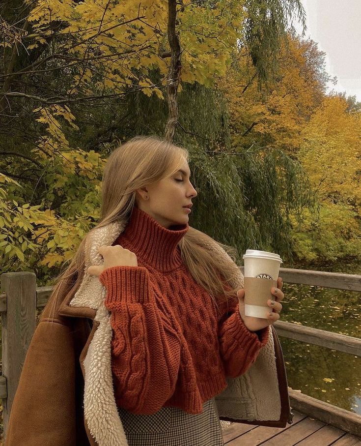 a woman holding a coffee cup while standing on a wooden bridge in the fall time