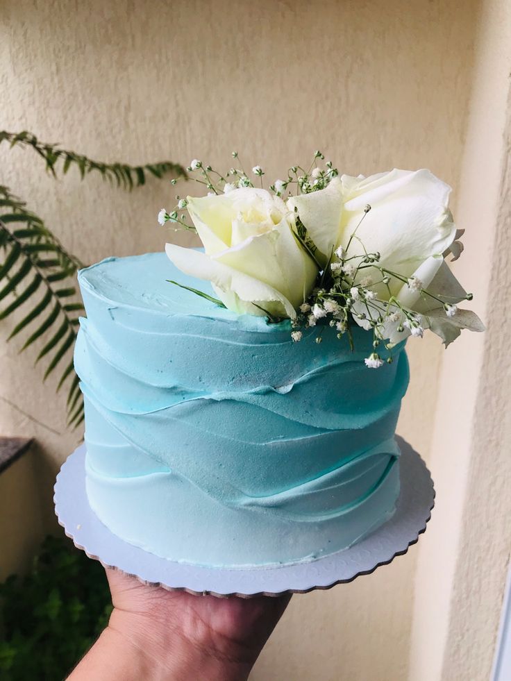 a hand holding a blue cake with white flowers on top and greenery in the middle