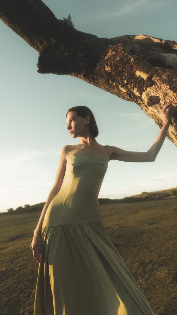 a woman in a long dress standing next to a tree branch with her hand on the trunk