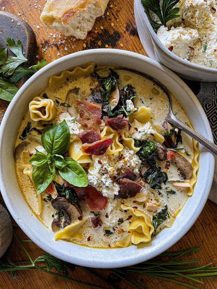 two bowls filled with pasta and spinach on top of a wooden table next to bread