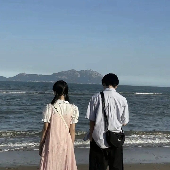 two people standing on the beach looking out at the ocean with mountains in the background