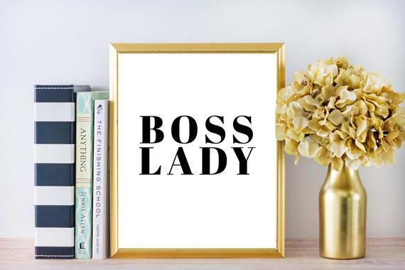 a gold vase with flowers next to a framed boss lady poster on a wooden table