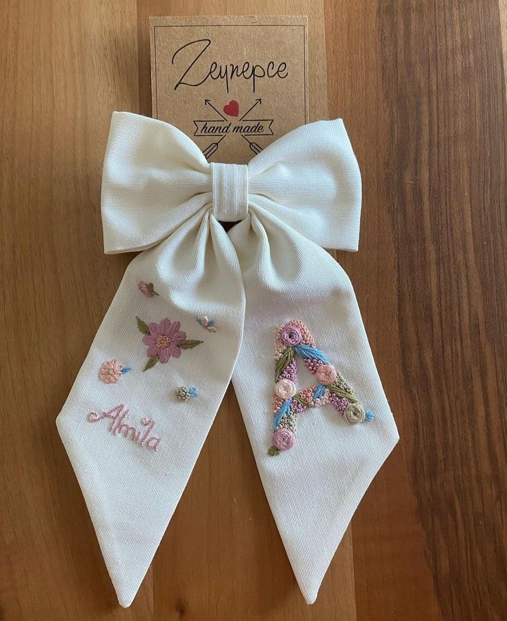 a white bow with embroidered letters and flowers on it, sitting on top of a wooden table