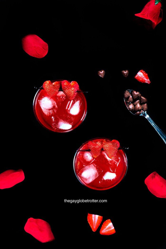 two glasses filled with liquid and strawberries on top of a black surface next to rose petals