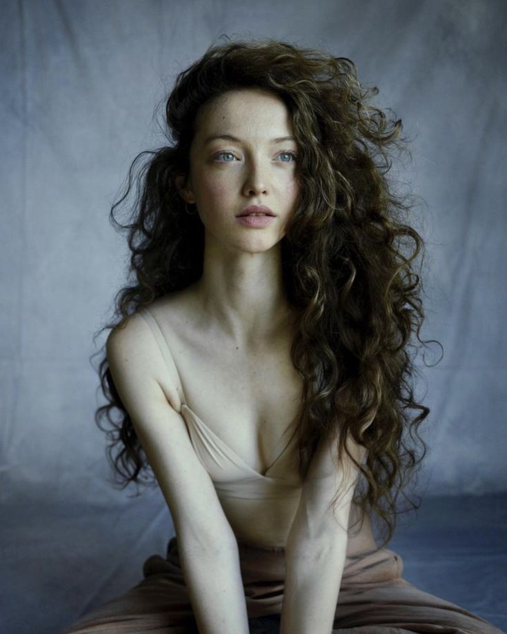 a woman with long hair sitting on the ground in front of a white backdrop and looking at the camera