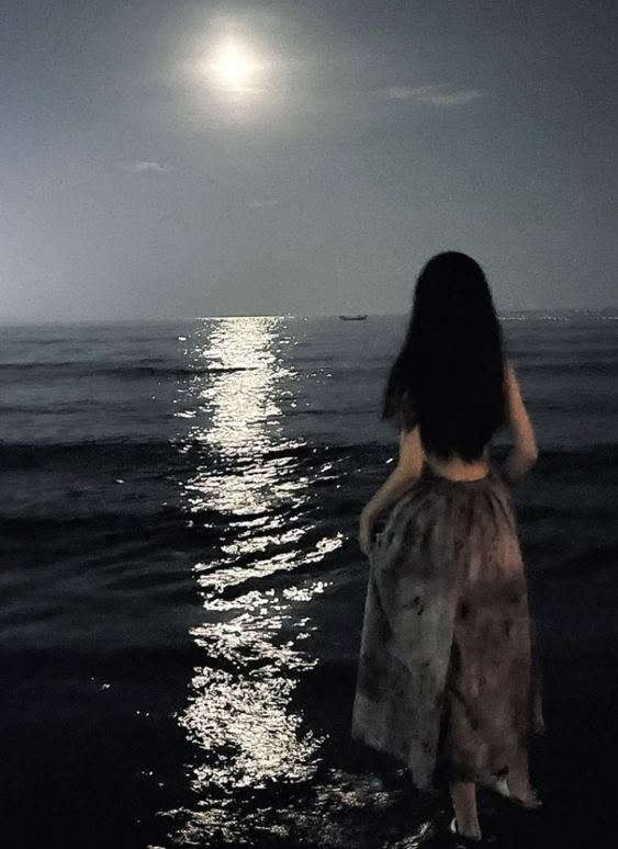 a woman is standing in the water looking at the moon over the ocean with her back to the camera