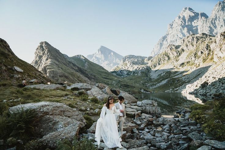 two women in white dresses are walking on rocks near the water and mountain peaks,