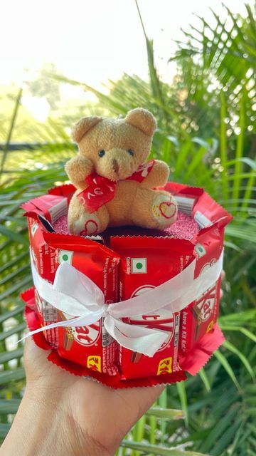 a hand holding a small teddy bear in a red box with candy bars on it