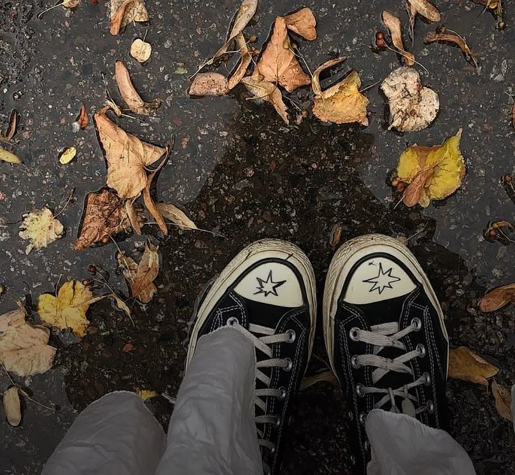 a person standing in front of leaves on the ground with their feet up and wearing converse shoes