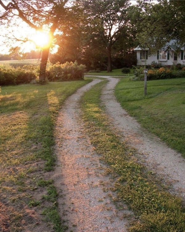 the sun is setting on a dirt path in front of a white house and trees