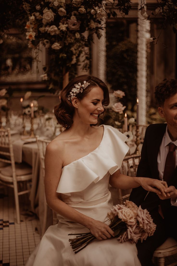 a bride and groom sitting at a table with flowers in their hair, holding each other's hands