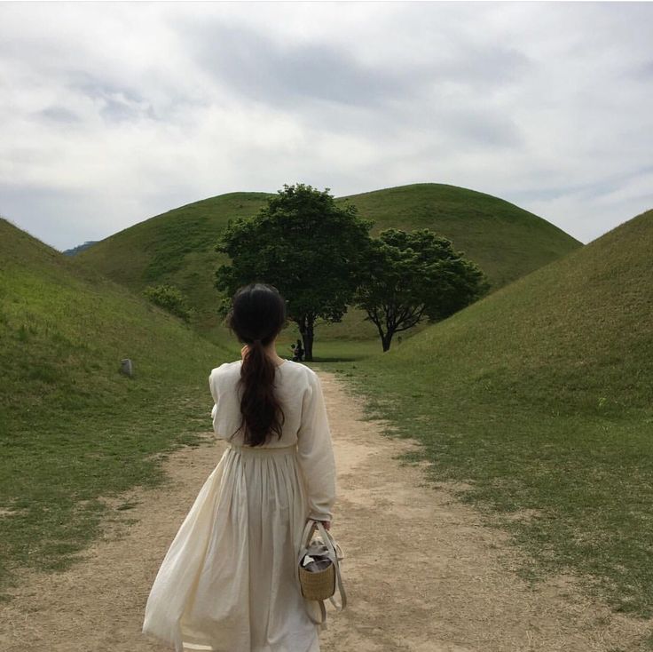 a woman in a white dress walking down a dirt road with trees on the hill behind her