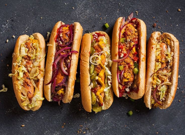 four hot dogs with various toppings lined up on a black surface, top view