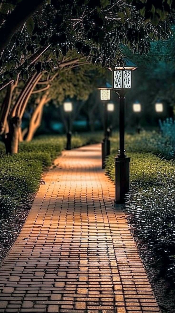 a brick path with two lamps on each side and trees in the background at night