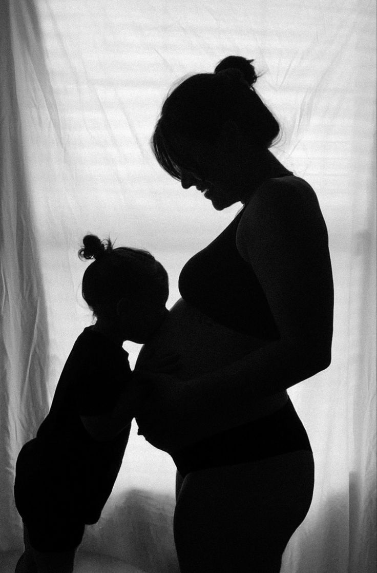 a pregnant woman holding a teddy bear in front of a window with white curtain behind her