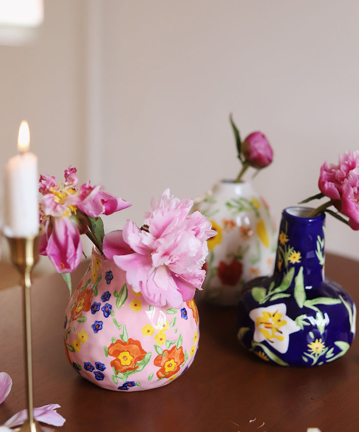 three colorful vases with flowers on a table next to a candle and some candlesticks