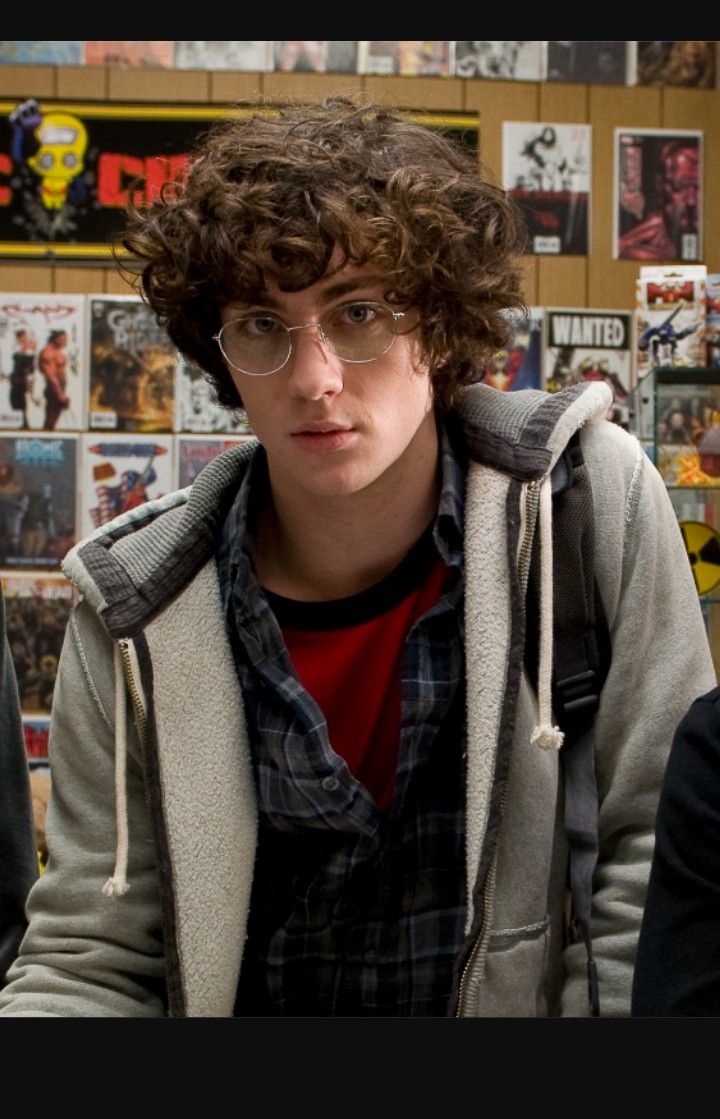 a young man with curly hair and glasses is looking at the camera while standing in front of a wall full of comic books