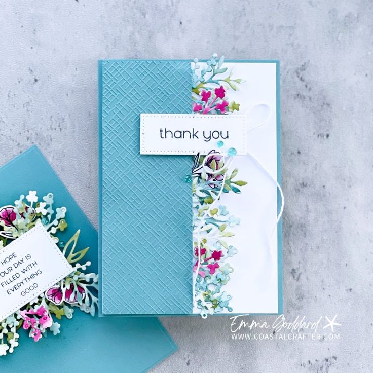 a thank you card with flowers on it next to a blue envelope and white tag