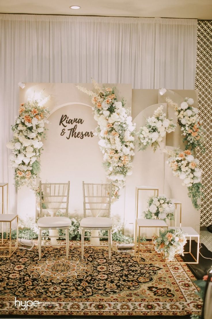 a wedding ceremony setup with white chairs and flowers on the back wall, in front of a floral backdrop